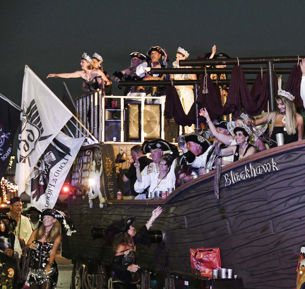 Parade float with people dressed as pirates onboard and walking throwing beads in a parade at night
